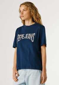 Młoda kobieta z falującymi włosami, ubrana w granatowy t-shirt "PEPE JEANS" oraz jasnoniebieskie jeansy, stojąca na tle jednolitego tła.