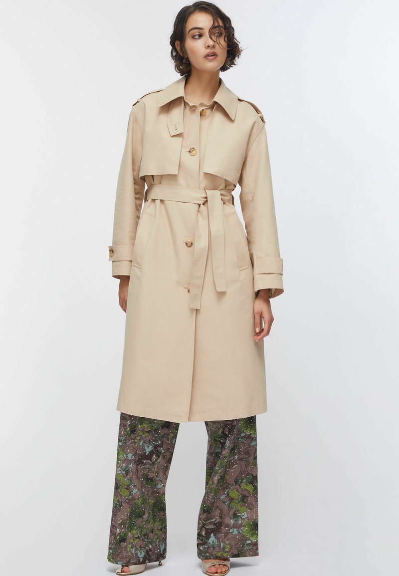 Kvinde iført en beige trenchcoat med bælte over blomstrede bukser med vide ben og åbne sko, stående mod en ensartet hvid baggrund.