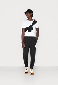T-shirt bianco in cotone, pantaloni della tuta neri con logo, cappello a secchiello nero e scarpe da ginnastica colorate. Outfit comodo e alla moda con uno stile a strati.