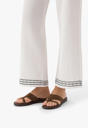 Stopy w brązowych zamszowych sandałach z pętelkami na palce w połączeniu z białymi spodniami z czarnymi, haftowanymi zakończeniami nogawek.