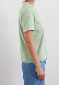 Nainen, jolla on vaaleanvihreä lyhythihainen polo-paita ja siniset farkut, seisoo sivuttain yksinkertaista valkoista taustaa vasten.