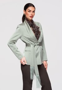 Femme portant un blazer vert menthe ceinturé avec une écharpe à franges et un foulard imprimé léopard, posant devant un fond clair uni.