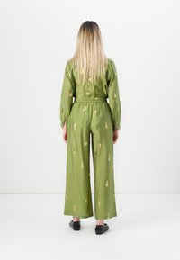 Zelen kombinezon iz tkanine z dolgimi rokavi, elastičnim pasom, širokimi nogami in vzorci z zlatimi listi. Obuti so črni ravni čevlji.