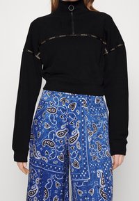 Sudadera negra recortada con cuello alto y media cremallera, que presenta cinta con logo dorado. Combinada con pantalones amplios de patrón paisley en azul.