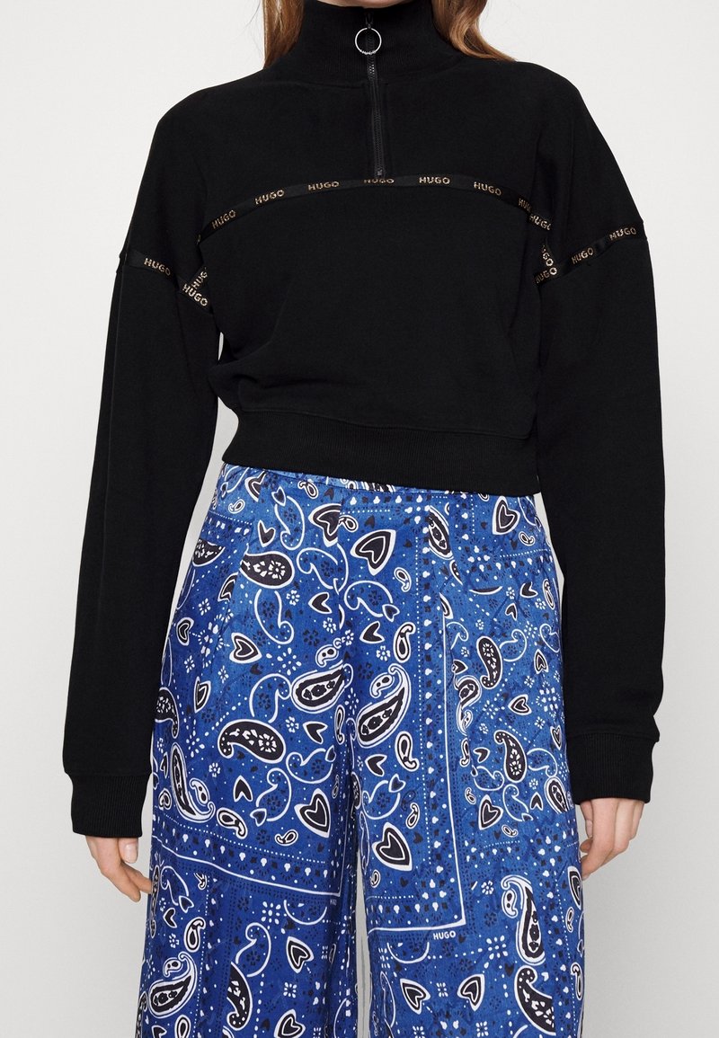 Sudadera negra recortada con cuello alto y media cremallera, que presenta cinta con logo dorado. Combinada con pantalones amplios de patrón paisley en azul.
