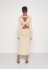 Robe en maille côtelée beige à manches longues, dos ouvert et détails croisés. Silhouette ajustée avec un ourlet arrivant à la cheville.