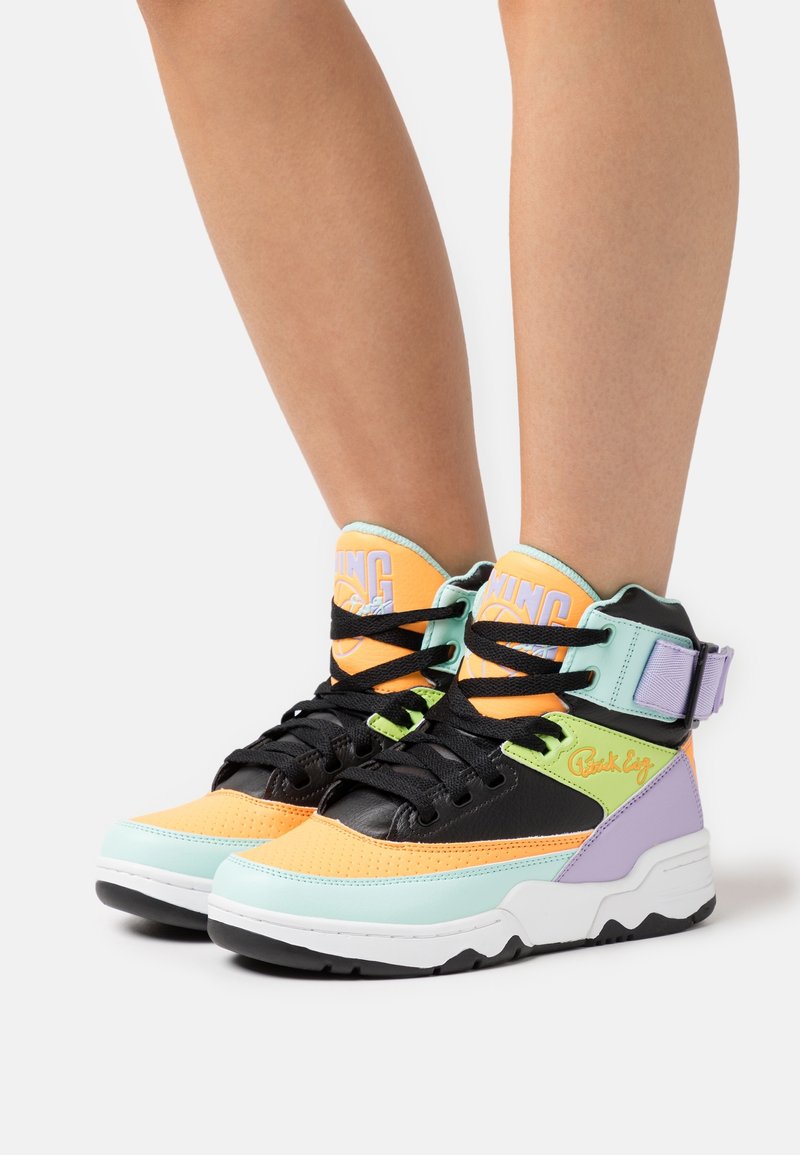 Baskets montantes avec des lacets noirs dans des couleurs pastel orange, bleu clair, vert et violet portées sur des jambes nues sur fond blanc.