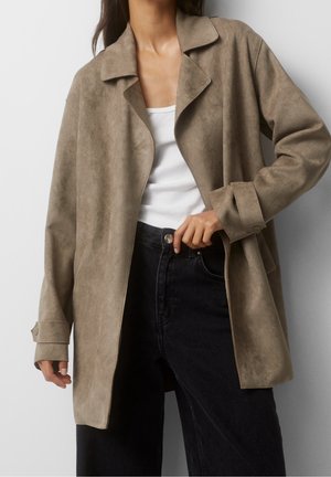 Femme portant une veste ouverte en daim beige sur un débardeur blanc et un jean noir taille haute, main ajustant le bouton du jean.