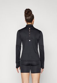 Maglietta sportiva nera a maniche lunghe con colletto alto, tessuto testurizzato e design a occhiello sul retro. Abbinata a pantaloni shorts neri.