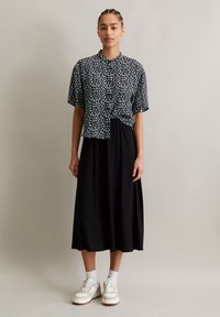 Schwarz-weiße gemusterte Bluse mit kurzen Ärmeln, kombiniert mit einem fließenden schwarzen Maxirock. Das Modell trägt weiße Sneakers und Knöchelsocken.