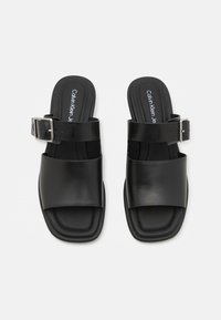 Sandales en cuir noir avec de larges brides, une forme carrée et une semelle texturée. Comprend une boucle en argent sur une des brides.