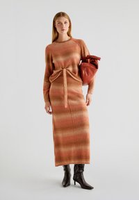 Robe en tricot à rayures orange et beige, présentant une taille nouée ; associée à un sac marron décontracté et des bottes noires jusqu'au genou.