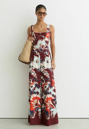 Kvinde iført en ærmeløs buksedragt med blomsterprint og vide ben, der holder en flettet mulepose og har store solbriller på foran en ensartet hvid væg.