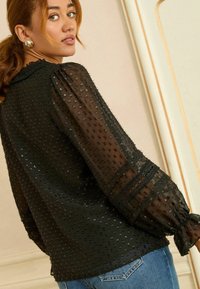 Melna caurspīdīga blūze ar tekstūru rotājumiem, pūkainām piedurknēm un mežģīņu akcentiem pie aprocēm. Kombinēta ar gaiši ziliem džinsu biksēm.