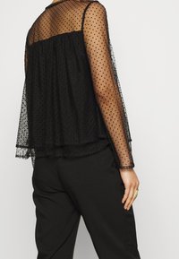 Svart genomskinlig blus med prickigt mönster, lagerdesign, långa ärmar och rundad halsringning, kombinerad med svarta byxor.