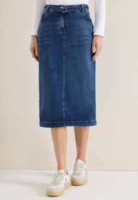 Femme portant une jupe en jean bleu foncé mi-longue avec une ceinture fine, des baskets blanches et un haut blanc à manches longues, debout sur un sol blanc.