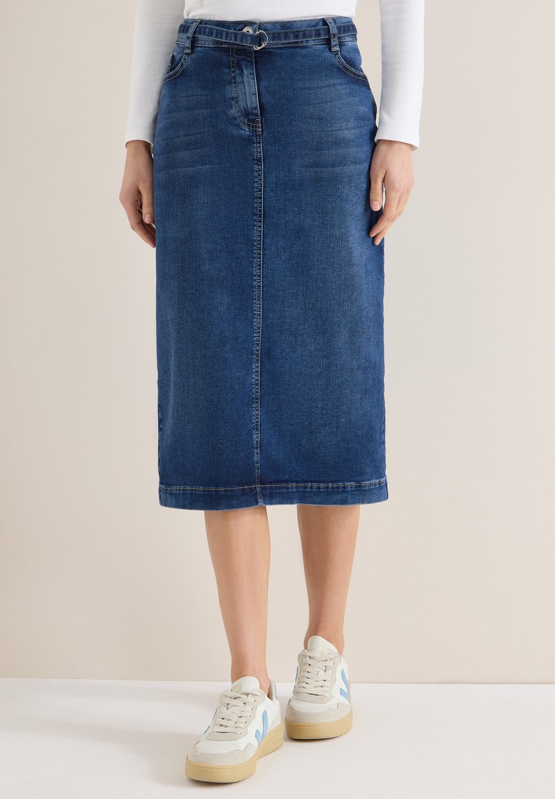 Femme portant une jupe en jean bleu foncé mi-longue avec une ceinture fine, des baskets blanches et un haut blanc à manches longues, debout sur un sol blanc.