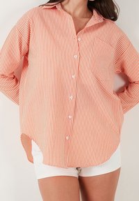 Chemise orange à rayures, boutonnée, manches longues, avec une poche sur le côté gauche et un ourlet arrondi. Fabriquée en tissu léger.