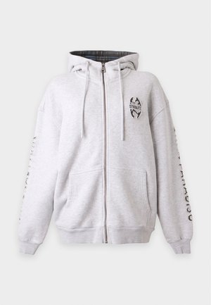 Junge Person mit beiger Kappe, weißem Hoodie, rot gestreiftem Hemd, hellblauer Hose und weißen Sneakers steht vor einem schlichten Hintergrund.