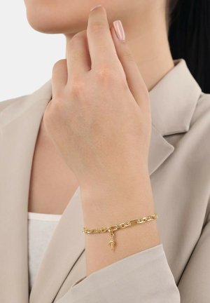 Poignet de femme portant un bracelet en chaîne dorée avec un petit charm en forme de palmier, vêtue d'un blazer beige et d'un haut blanc.