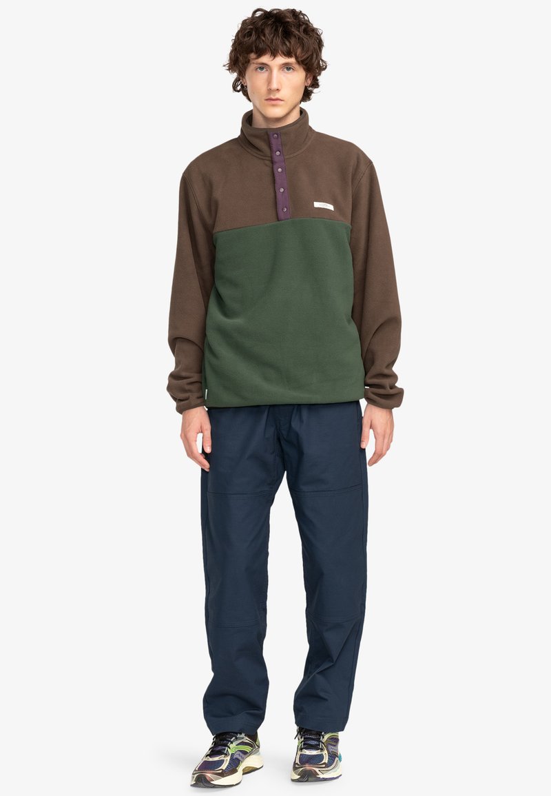 Pull en polaire marron avec une partie inférieure verte, doté d'un col montant, d'accents en bouton violet et d'un petit logo. Associé à un pantalon bleu marine.