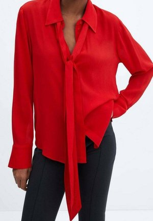 Blouse rouge à manches longues avec col et nœud sur le devant, portée avec un pantalon noir ajusté, portée par une personne avec une main sur la hanche.