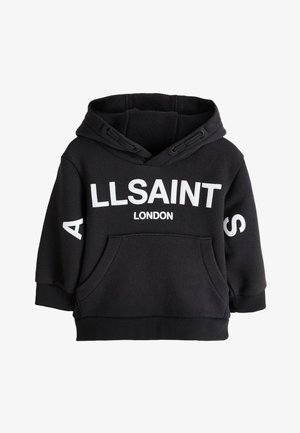 Smilende barn med flettet hår iført en sort AllSaints London hættetrøje og sorte bukser, stående mod en ensfarvet lys baggrund.