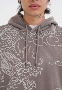 Felpa grigia con intricate ricami bianchi di drago, cappuccio con lacci e una texture morbida. Nota per i suoi dettagli di design.