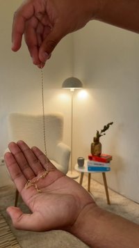 Colar de corrente de ouro segurado numa mão, apresentando elos intrincados. Ao fundo, uma cadeira estofada, uma lâmpada e livros numa mesa lateral.