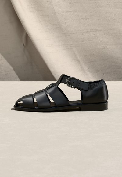 Sandale plate en cuir noir avec talon fermé, trois brides horizontales, une bride à boucle à la cheville, et design à bout ouvert.