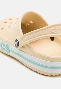 Beżowe Crocs z paskiem z tyłu, z otworami wentylacyjnymi i teksturowaną wkładką. Akcentowane białym i miętowym paskiem.