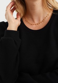 Svart strukturerad tröja med rund halsringning, i kombination med ett elegant guldhalsband som har små, spetsiga detaljer och ett fint kedja.