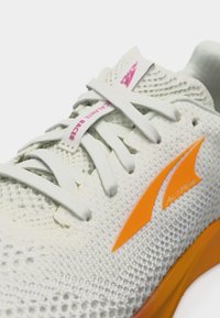 Άσπρο πλεκτό αθλητικό παπούτσι με άσπρα κορδόνια, ροζ ετικέτα "Volcanic Pacer" και πορτοκαλί λογότυπο Altra στο πλάι και στη σόλα.