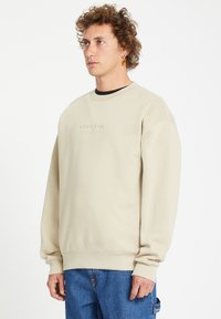 Beige sweatshirt with a crew neck and ribbed cuffs. Features embroidered "VOLCOM" logo at the chest. Paired with blue denim jeans.