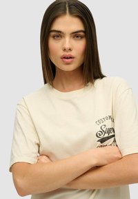 Camiseta de algodón beis de mangas cortas, con un estampado gráfico negro en el lado izquierdo del pecho. Corte recto y textura suave.