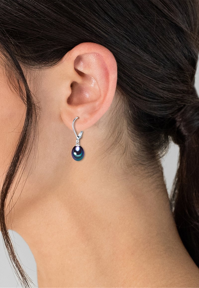 Primo piano dell’orecchio di una donna che indossa un orecchino ad amo in argento con una goccia di perla scura iridescente, capelli scuri raccolti.