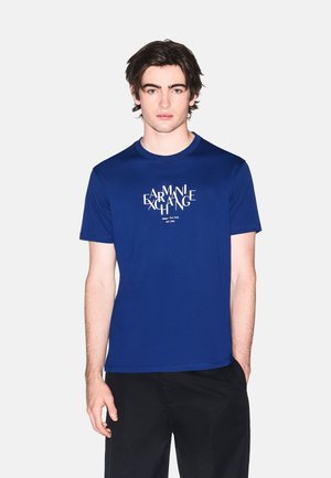 Giovane uomo con capelli scuri che indossa una t-shirt blu Armani Exchange con logo bianco, abbinata a pantaloni neri, in piedi davanti a uno sfondo semplice.