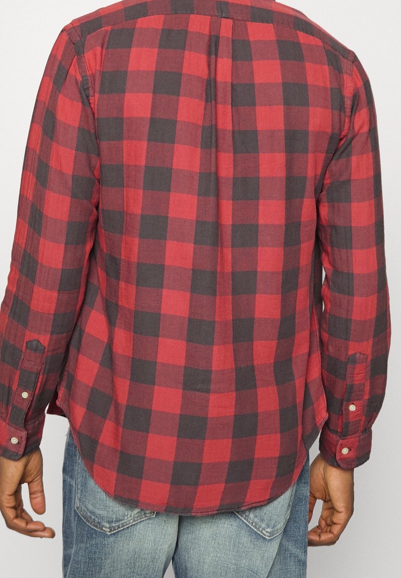 Homme portant une chemise à carreaux rouge et noire à manches longues et un jean bleu, vu de dos avec les bras détendus le long du corps.