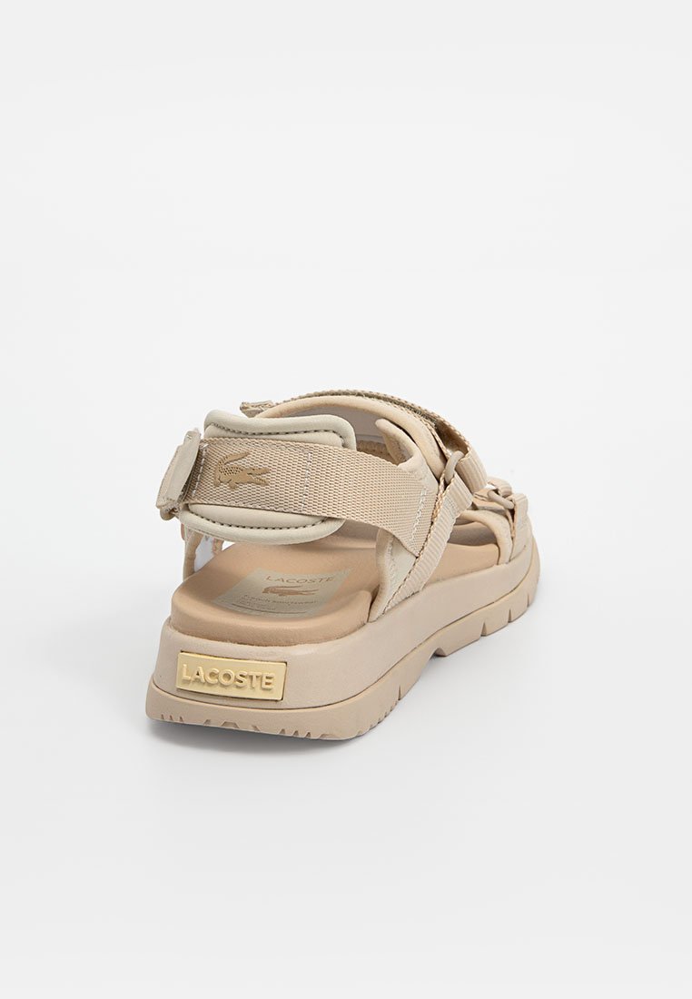 Sandale beige avec des sangles ajustables, matériau texturé et semelle épaisse. Présente un accent en logo doré sur le talon et un design incurvé.