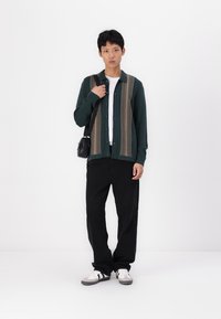 Cardigan verde con motivo a righe verticali, t-shirt bianca, pantaloni neri a vestibilità ampia e sneakers grigie con dettagli neri. Borsa a tracolla indossata.