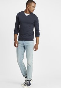Maglione blu navy a scollo a V in tessuto a maglia, abbinato a jeans azzurri con risvolto e sneakers blu navy alte, su uno sfondo neutro.