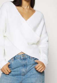 Weißer, gestrickter Wickelpullover mit V-Ausschnitt, der eine gerippte Struktur und überschnittene Schultern aufweist, kombiniert mit hoch taillierter blauer Jeans.