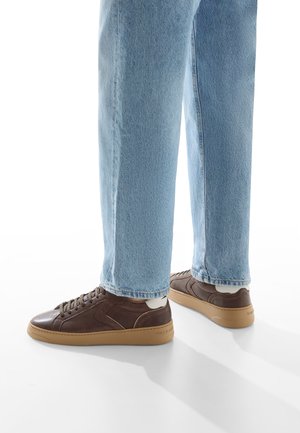 Sneaker in pelle marrone con finitura testurizzata e design con lacci, abbinati a jeans blu chiaro a gamba dritta. Suola in gomma e accenti a contrasto.