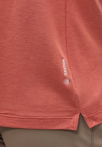 Κοντινή λήψη της βάσης ενός πουκαμίσου από υφασμάτινο, ροζ-σολομού χρώμα με μικρό λογότυπο "DRYTON" πάνω από μια πλαϊνή σκίσιμο πάνω από καφέ παντελόνι.