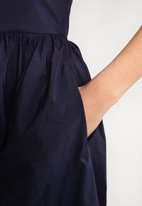 Dunkelblaues Kleid mit strukturiertem Stoff, gerafftem Bund und einer Seitentasche, in die eine Hand teilweise eingesteckt ist, um die Silhouette des Kleides zu zeigen.