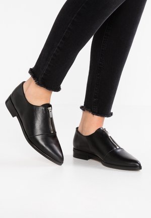 Pieds chaussés de chaussures en cuir noir à bout pointu avec fermetures éclair à l'avant, associés à un jean skinny noir à ourlet effiloché sur fond blanc.