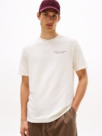 Balta kokvilnas t-krekls ar īsām piedurknēm un apaļu kakla izgriezumu, uz kura ir drukāts "TOMMY HILFIGER Established 1985" pelēkā krāsā. Brūni corduroy bikses.