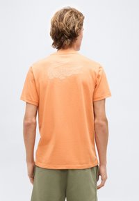 Νεαρός άνδρας με ανοιχτά καστανά μαλλιά που φοράει πορτοκαλί μπλούζα με λευκό σχέδιο κροκόδειλου στο πάνω μέρος της πλάτης, συνδυασμένη με χακί σορτς.