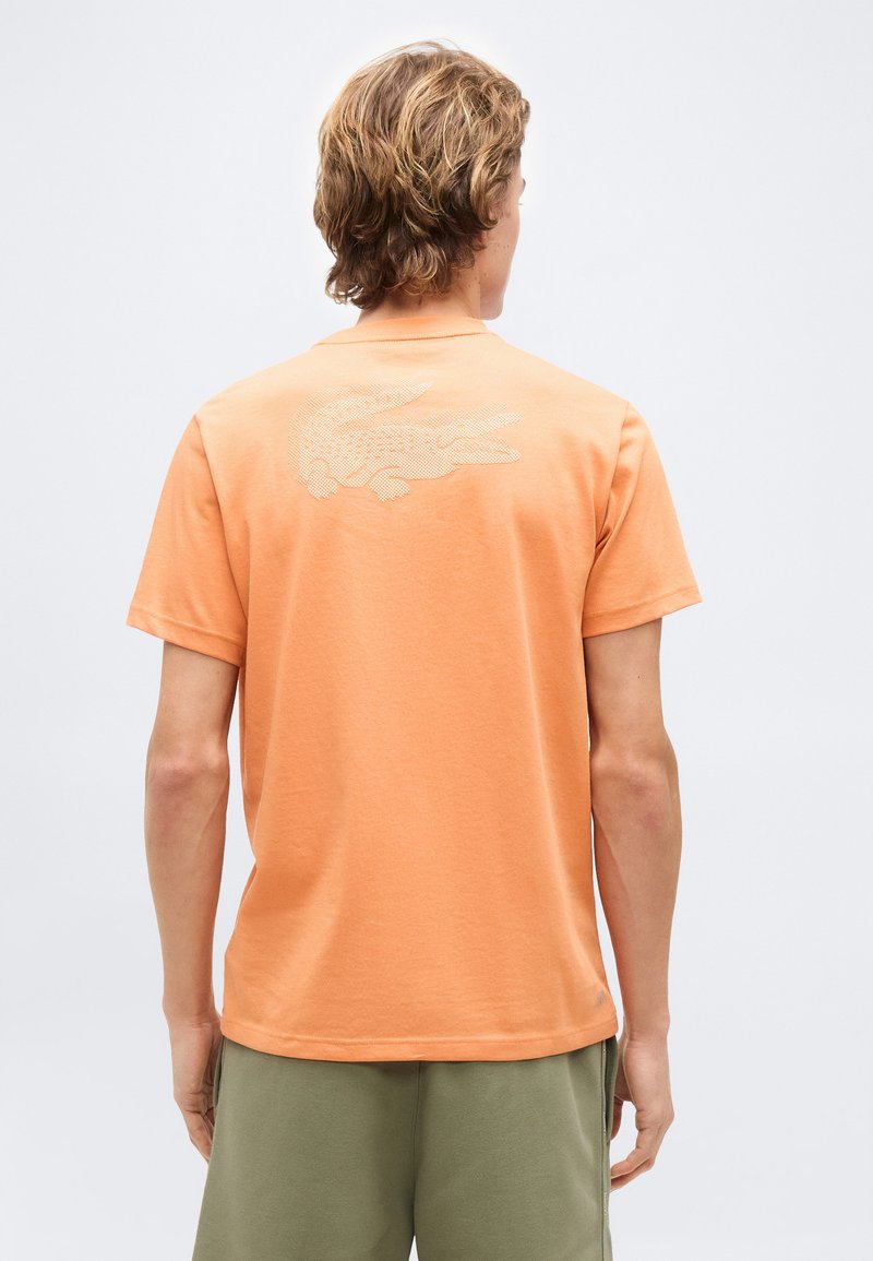 Νεαρός άνδρας με ανοιχτά καστανά μαλλιά που φοράει πορτοκαλί μπλούζα με λευκό σχέδιο κροκόδειλου στο πάνω μέρος της πλάτης, συνδυασμένη με χακί σορτς.