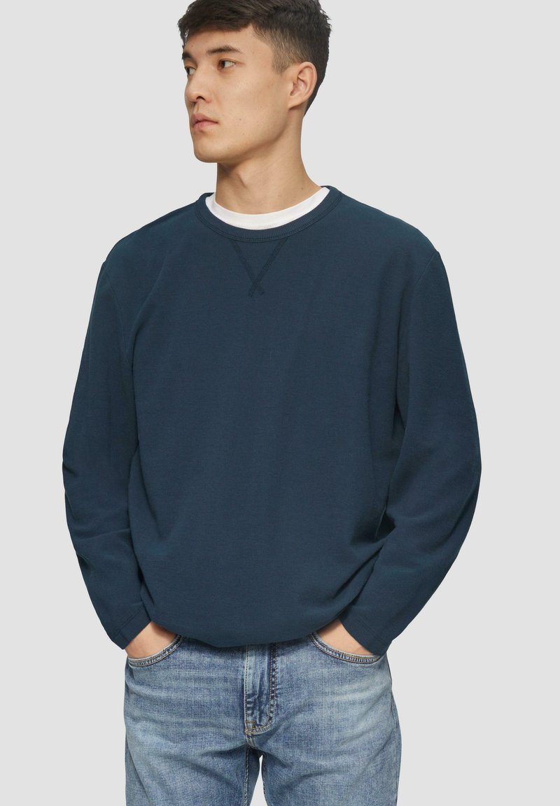 Dunkelblaues Sweatshirt mit Rundhalsausschnitt, langen Ärmeln und strukturiertem Stoff. Getragen mit hellblauen Jeans, Hände in den Taschen, minimalistisches Design.